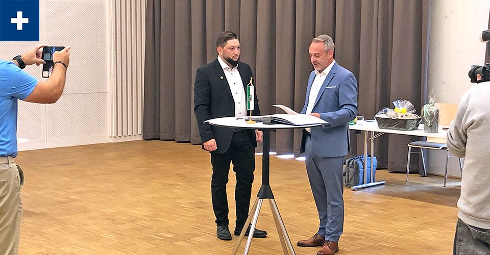 Zwei Männer in Anzügen stehen bei einem Tisch mit einer Flagge und Papieren, einer liest, während der andere zuhört. Eine Person macht im Hintergrund ein Foto.