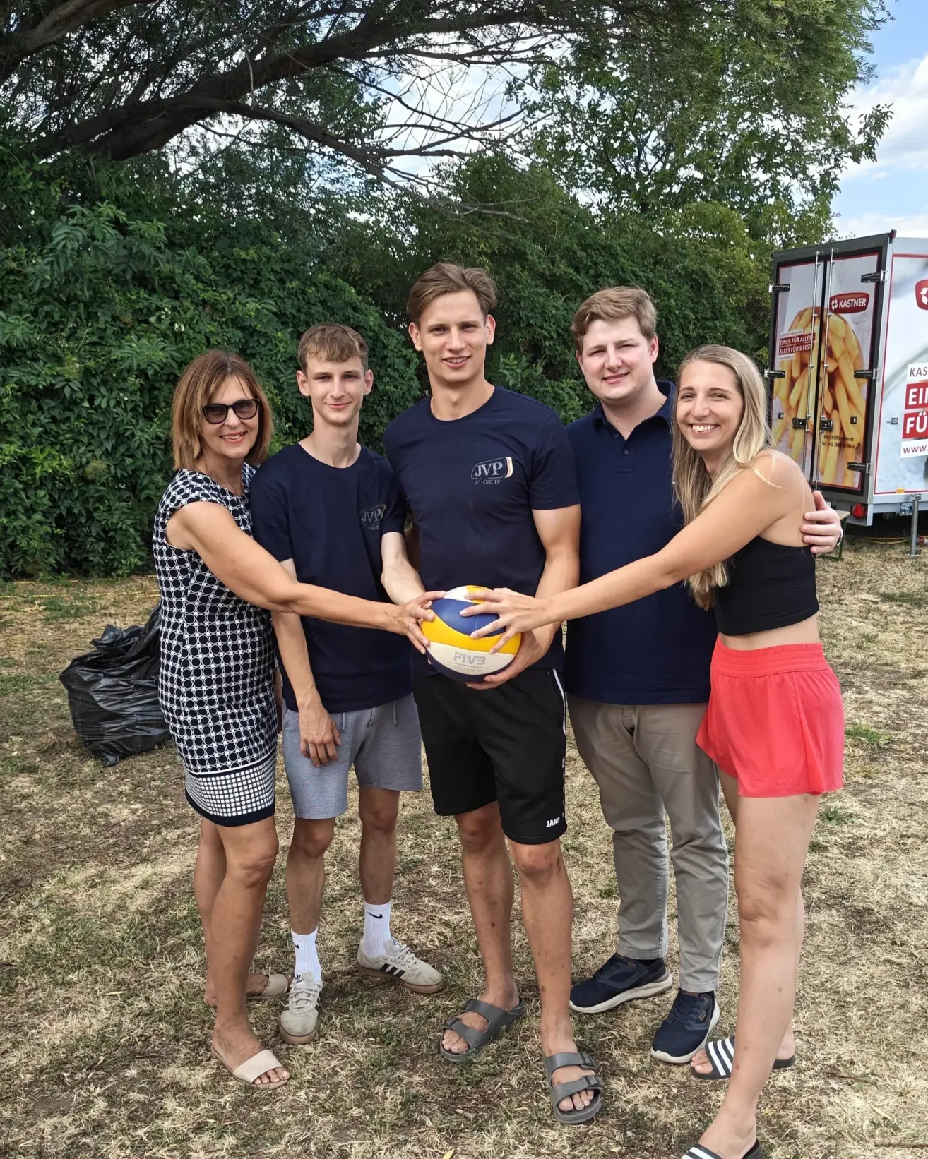 Eine Gruppe von fünf Personen, darunter zwei Frauen und drei Männer, posiert für ein Foto, während sie einen Volleyball halten. Sie stehen in einem Außenbereich mit einem LKW und Bäumen im Hintergrund.