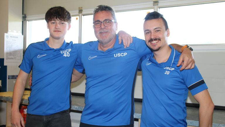 Three men in blue polo shirts stand together, smiling. The man in the middle has USCp 40 on his shirt, while the others have USCp on theirs. They are in a room with a window behind them.