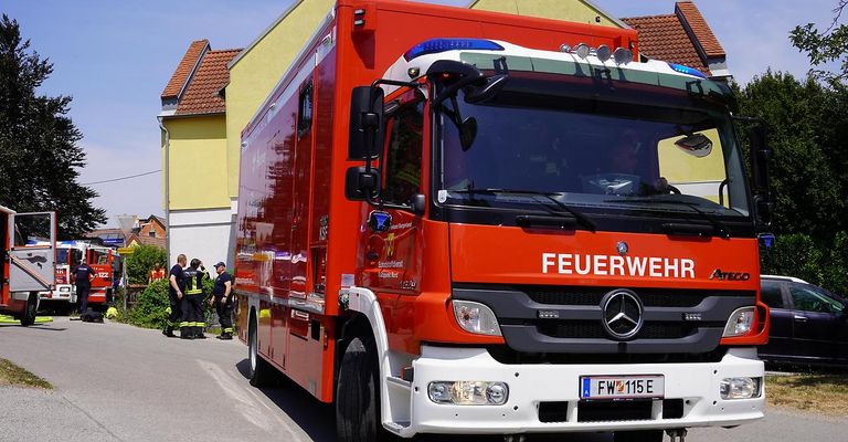 A red fire truck with the word 'Feuerwehr' on the front is parked on the side of the road with firefighters standing nearby.