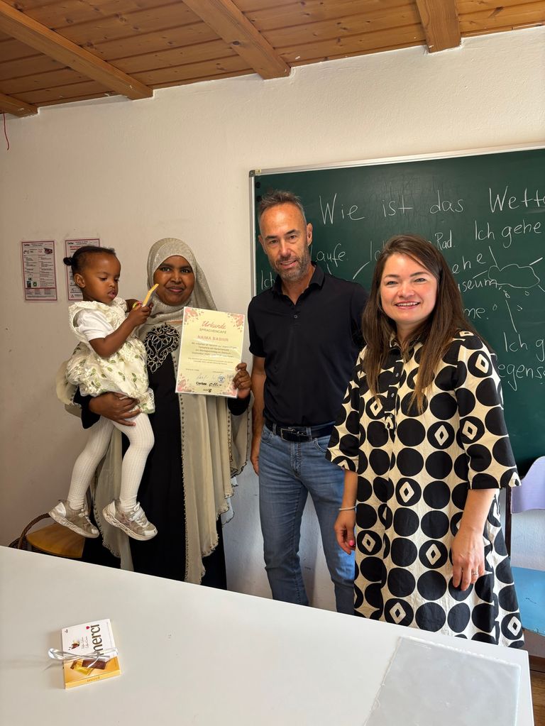 Eine Familie steht vor einer Tafel mit einem kleinen Mädchen, das ein Zertifikat hält und lächelt. Der Mann und die Frau neben ihr lächeln ebenfalls.