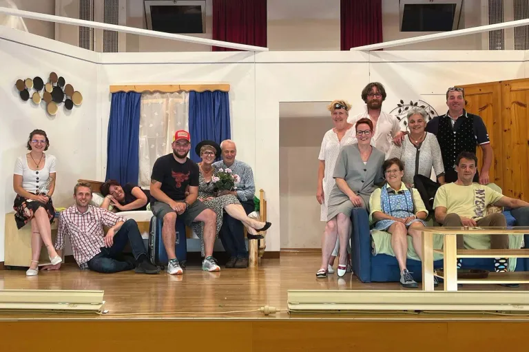 Eine Gruppe von Menschen posiert für ein Foto auf einer Bühne, einige sitzen auf einem Bett und andere stehen. Sie sind in verschiedenen Outfits gekleidet.