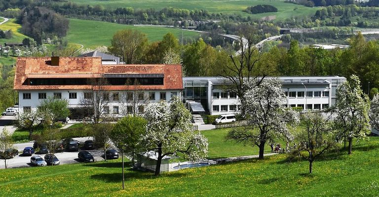 Luftaufnahme eines modernen Gebäudes mit rotem Dach, umgeben von Bäumen und einem grünen Feld. Vor dem Gebäude sind mehrere Autos geparkt, und eine Brücke ist im Hintergrund zu sehen.