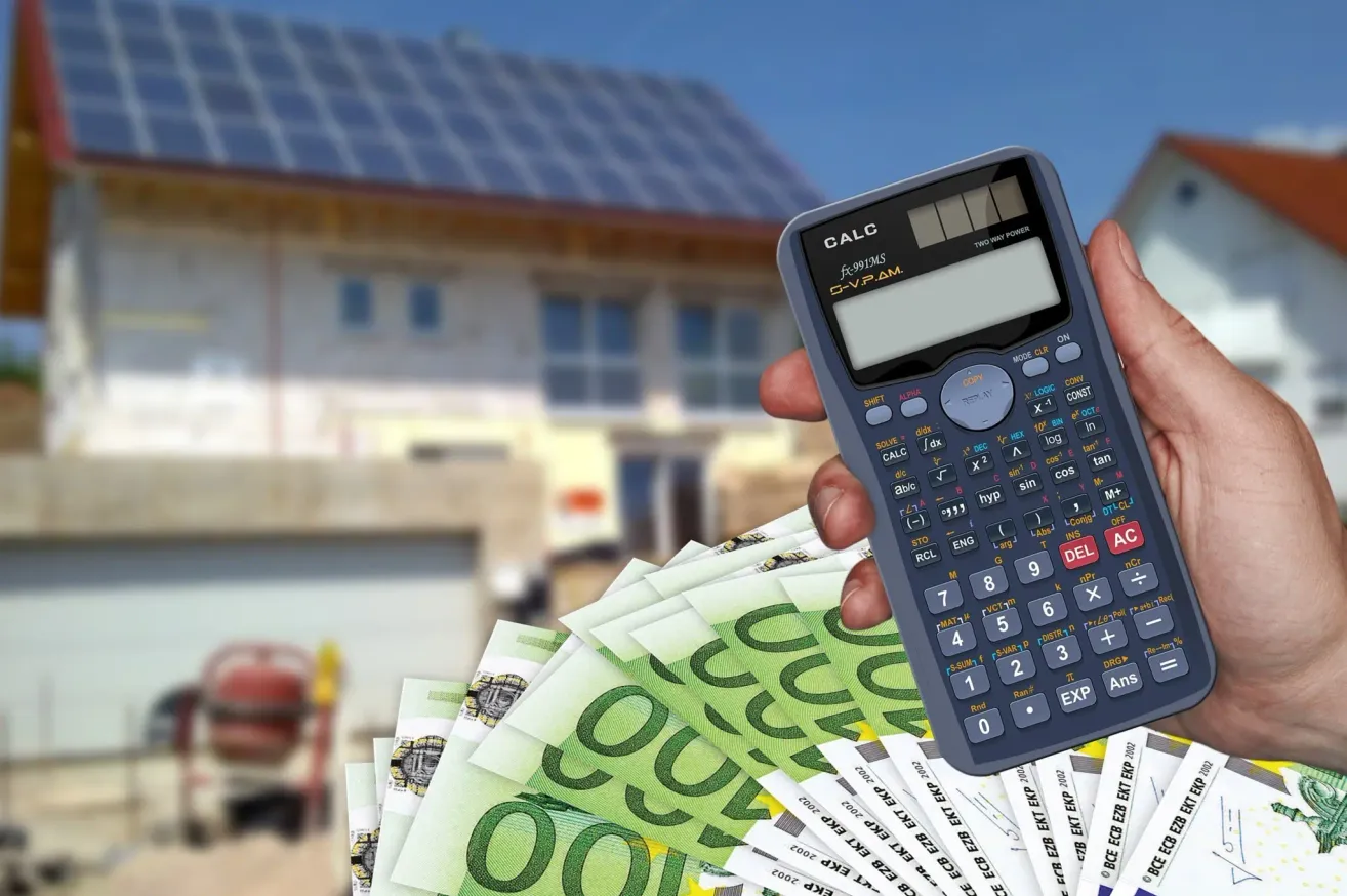 Eine Hand hält einen Taschenrechner über einem Haufen Geld mit einem Haus mit Solarpanelen im Hintergrund.