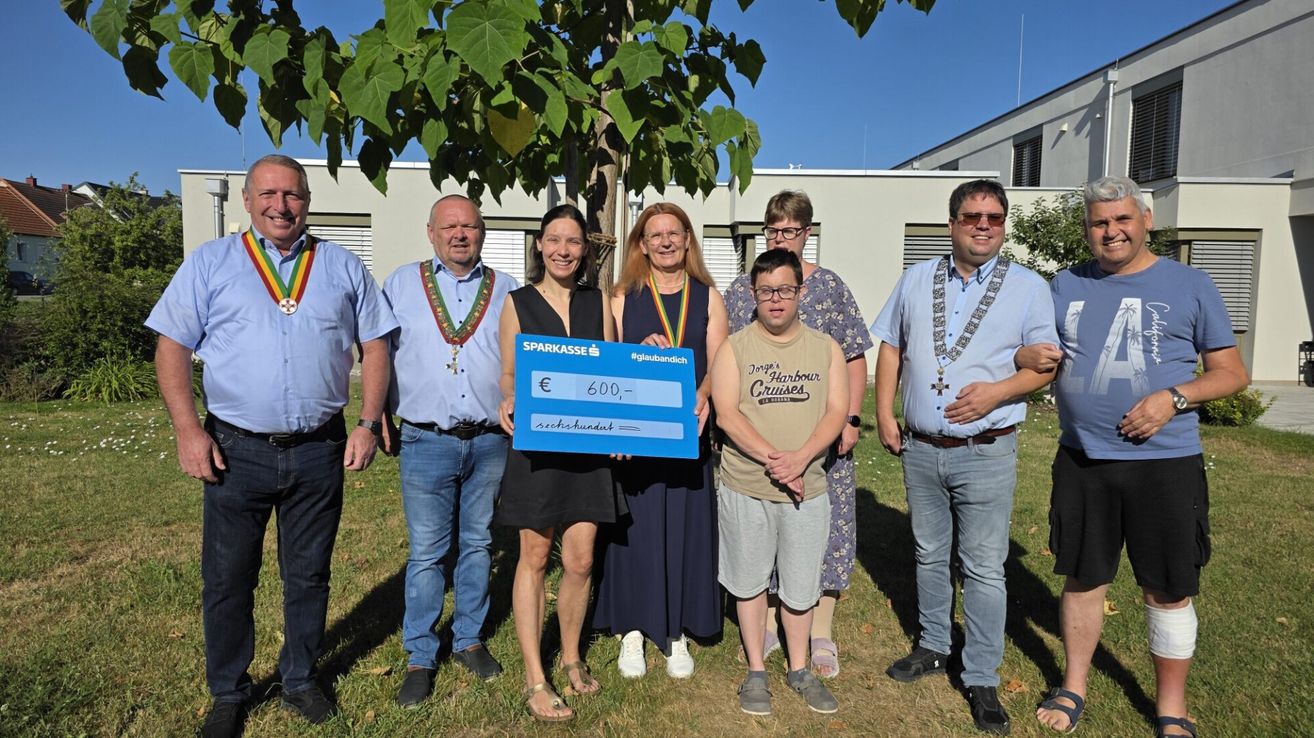 A group of people stand under a tree holding a large blue check. They are all smiling and appear to be posing for a photo.
