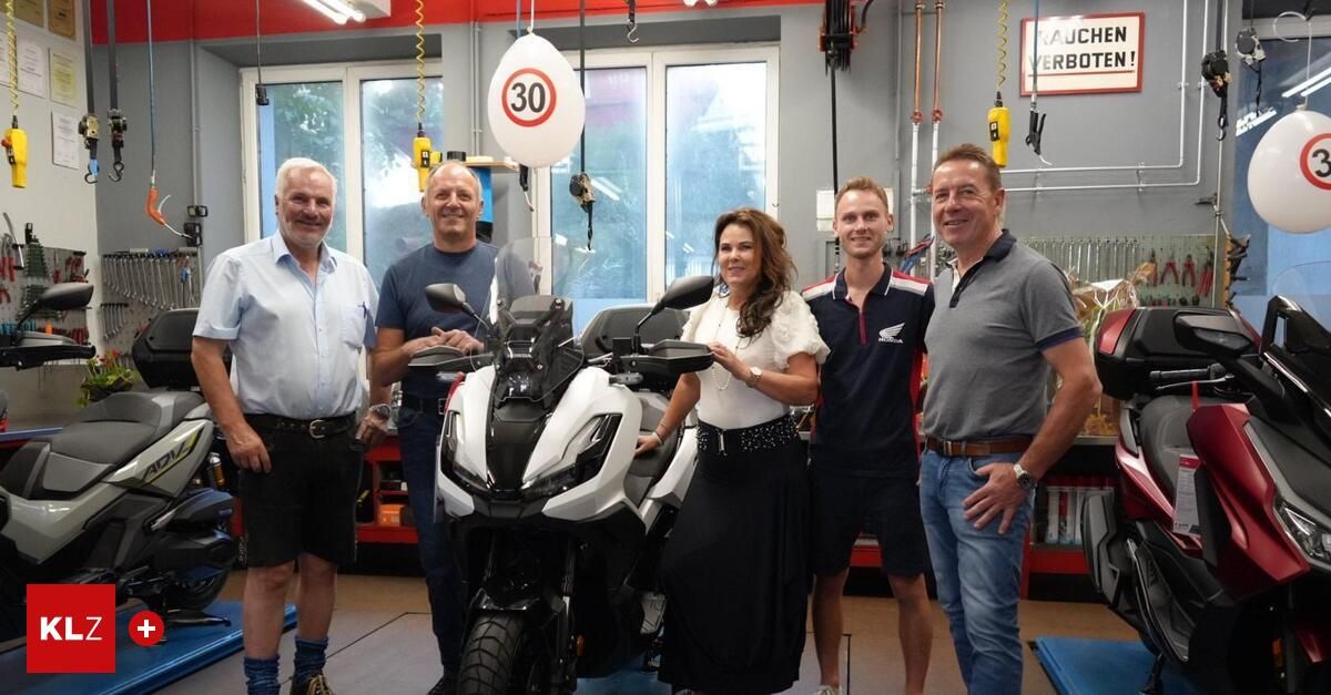 Fünf Personen stehen in einer Garage, lächeln und posieren für ein Foto. Eine Frau in der Mitte hält das Lenkrad eines weißen Motorrads. Dahinter befindet sich ein Regal mit verschiedenen Gegenständen, und ein weißer Ballon mit der Nummer 30 hängt von der Decke.