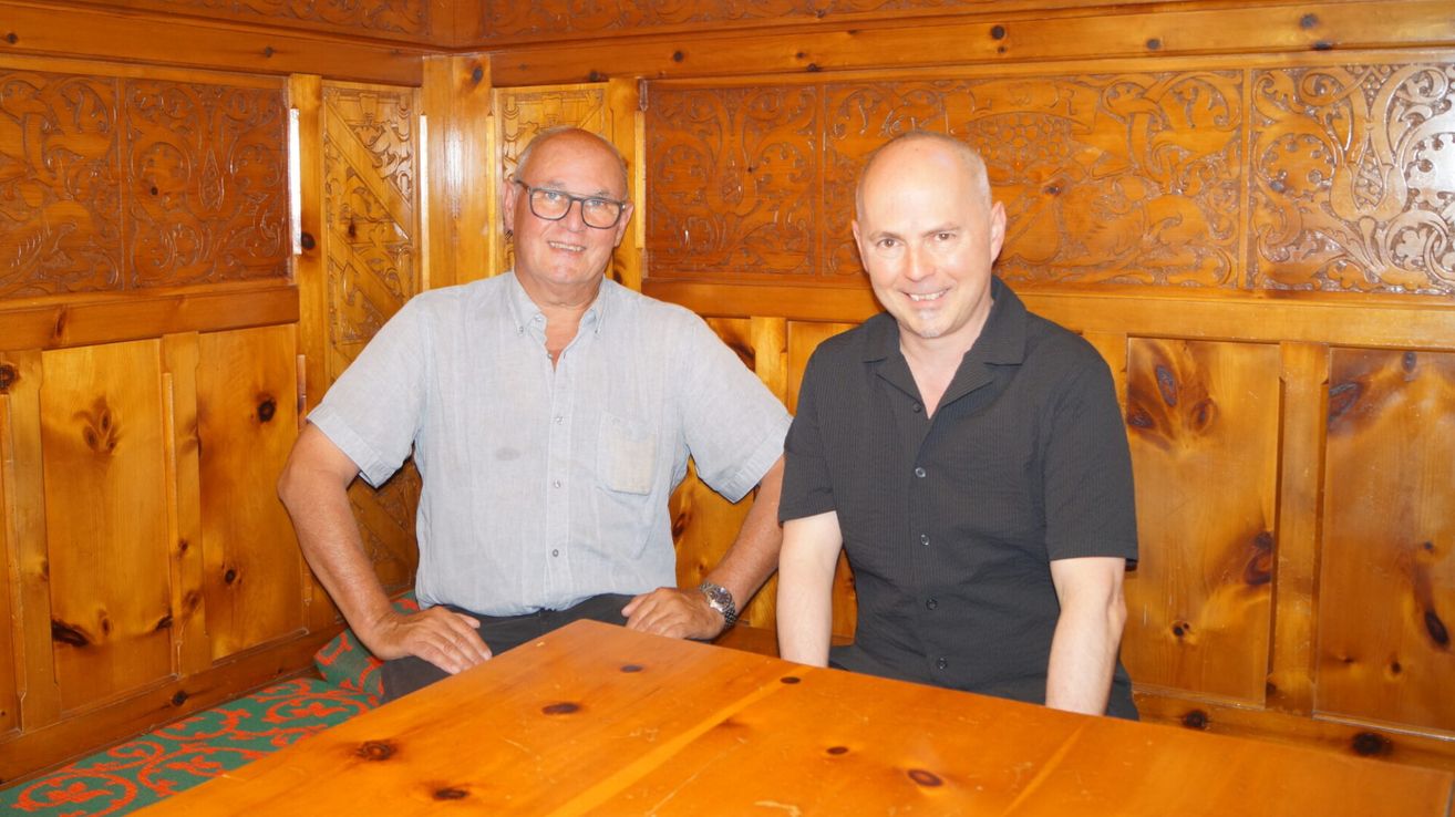 Two men sitting at a wooden table, smiling, with a carved wooden wall in the background.
