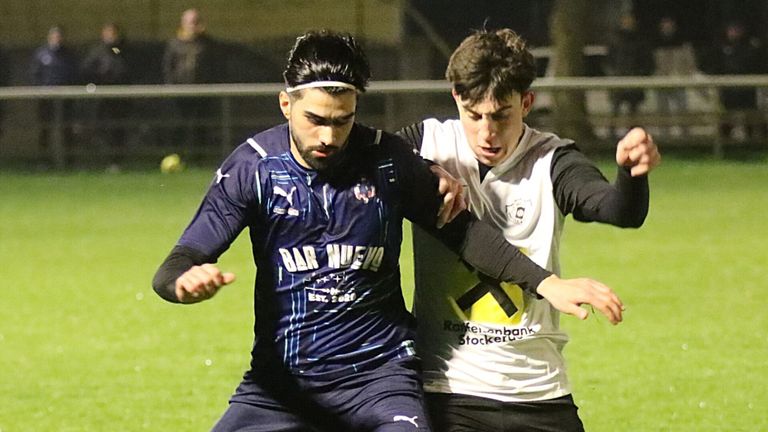 Two soccer players are engaged in a physical duel on the field. The player in blue is wearing a headband and has 'Bar Nuevo' on his jersey. The player in white has a logo on his jersey.
