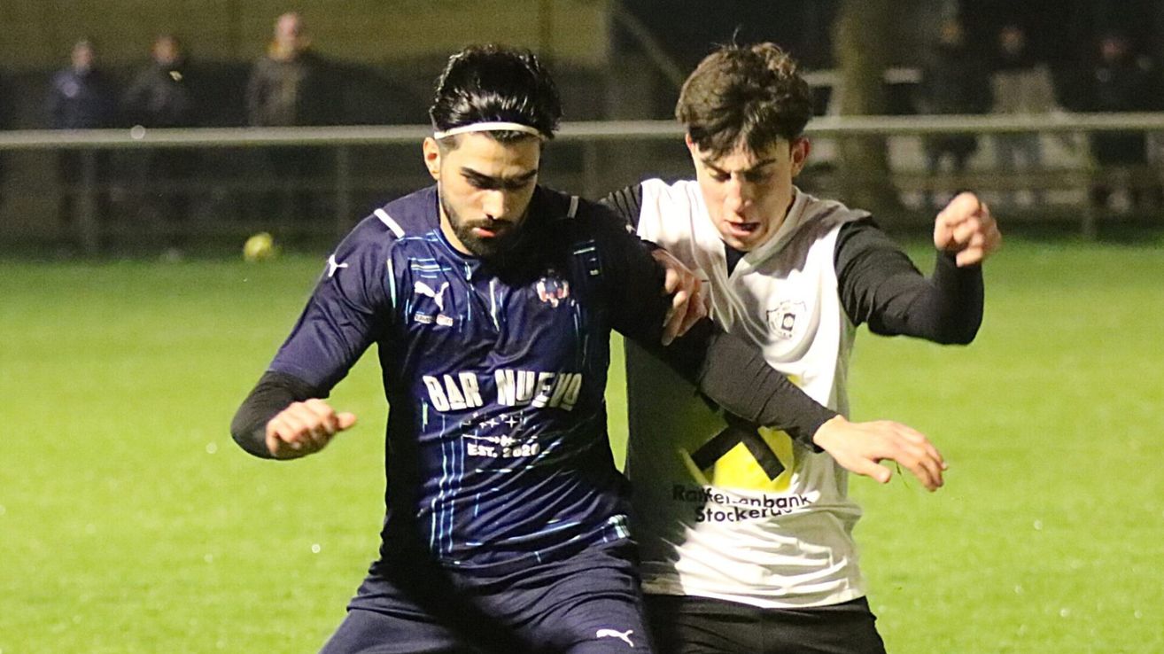Two soccer players are engaged in a physical duel on the field. The player in blue is wearing a headband and has 'Bar Nuevo' on his jersey. The player in white has a logo on his jersey.