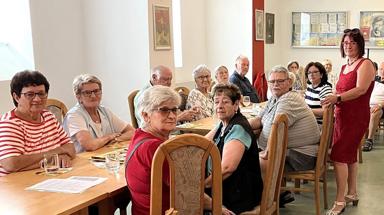 A group of elderly people sitting around a table in a dining hall, some wearing glasses and smiling.