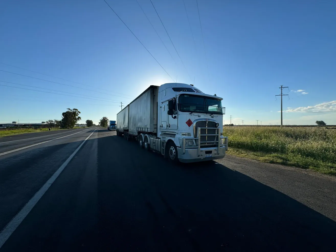 A white semi-truck is driving on a highway with a clear blue sky and a field on the side.