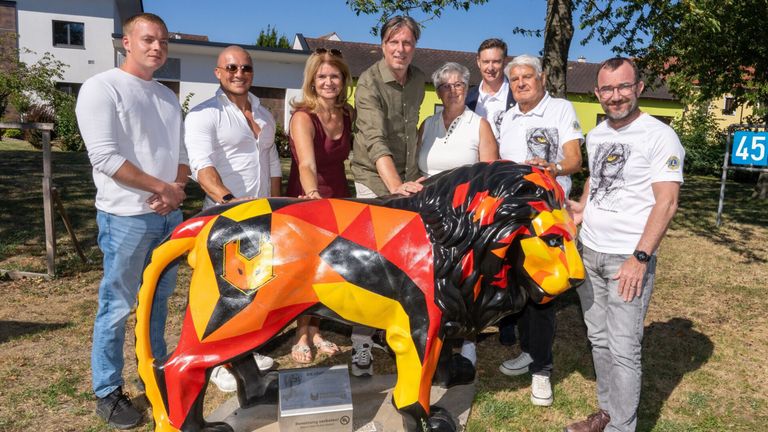 A group of people are posing with a colorful lion statue in a park. The statue is painted in red, yellow, and black. The people are smiling and seem to be posing for a photo.