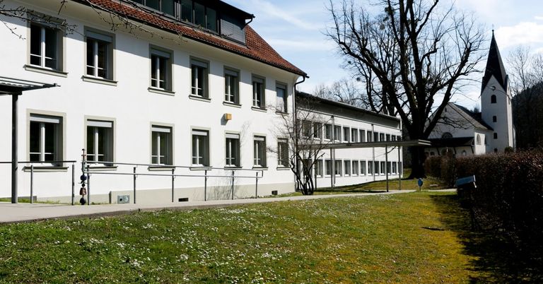 Ein weißes Gebäude mit vielen Fenstern und einem roten Dach ist von einem Grasbereich mit einem Baum und einem Gehweg umgeben.