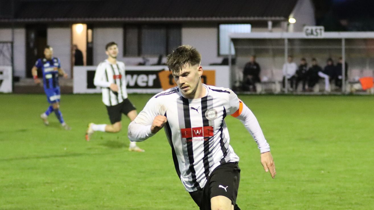 A soccer player in a white and black striped jersey is running on the field, looking ahead. Another player runs behind him. In the background, there is a building with a banner.