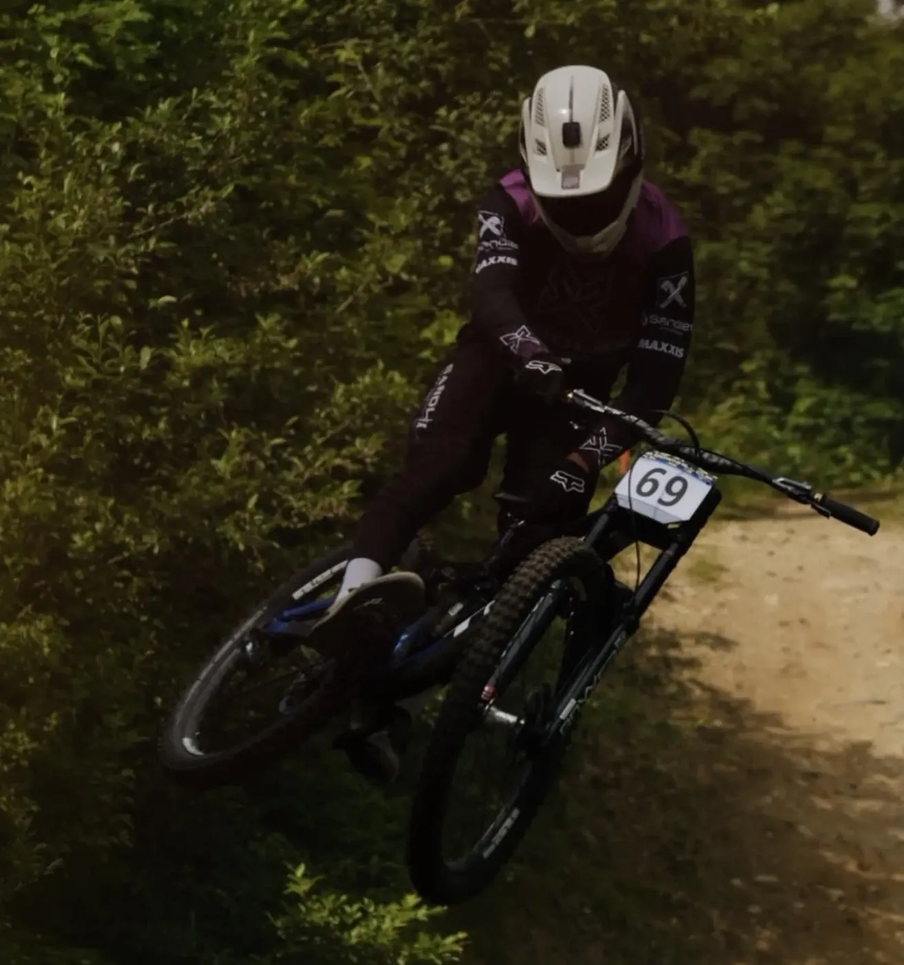 Ein Mountainbiker mit weißem Helm und Trikot mit der Nummer 69 fährt ein Fahrrad in der Luft auf einem mit grünem Laub umgebenen Erdweg.