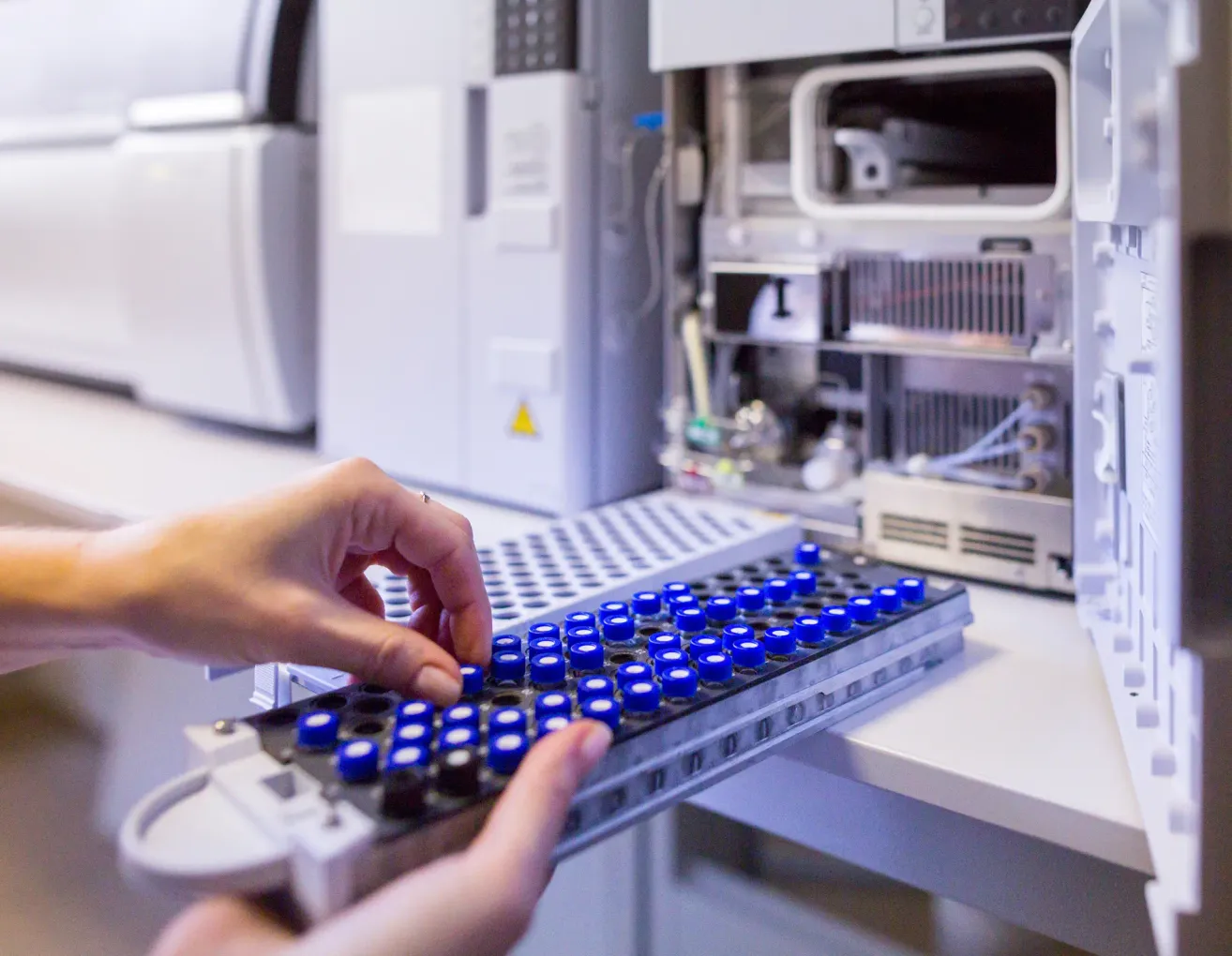 A person is handling a tray with blue vials in a laboratory setting, with lab equipment visible in the background.