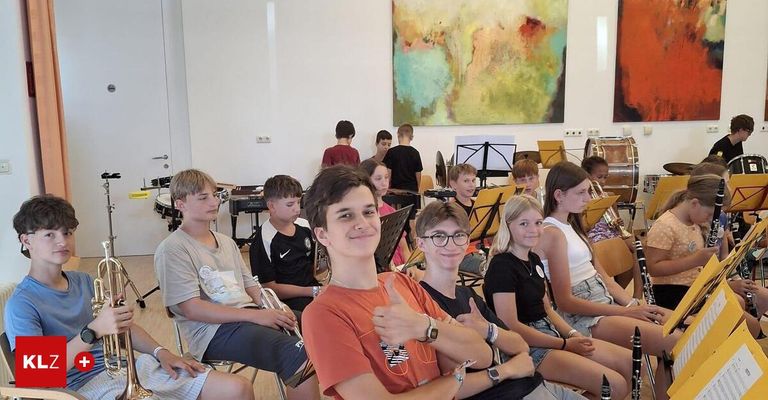 A group of young musicians, some holding instruments, sit in chairs in a music room with a colorful painting on the wall.