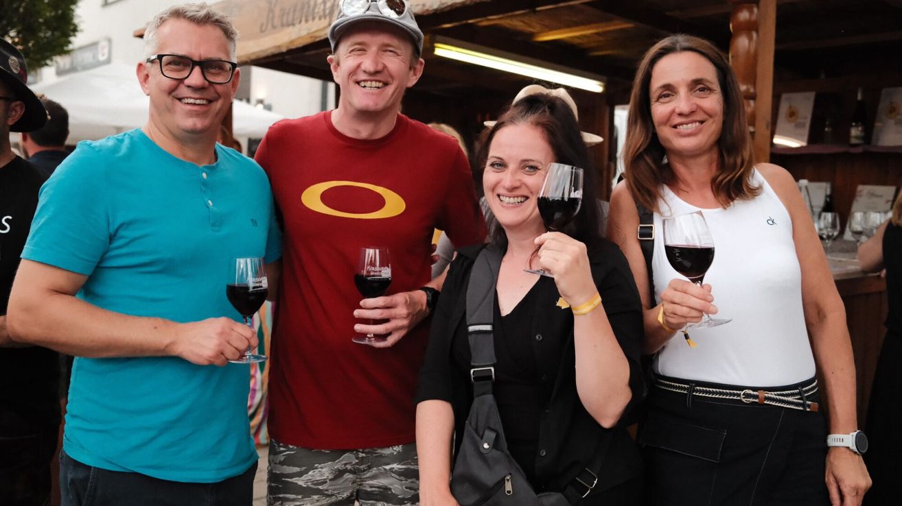 Vier Menschen, die Wein in Gläsern halten und lächeln, zwei Frauen und zwei Männer, stehen in einer festlichen Umgebung.