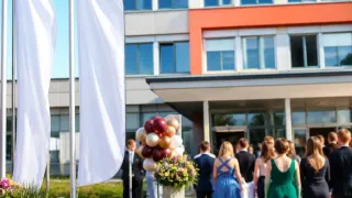 A group of people dressed in formal attire are gathered outside a modern building with large windows. A bouquet of flowers and a cluster of balloons are in the foreground.