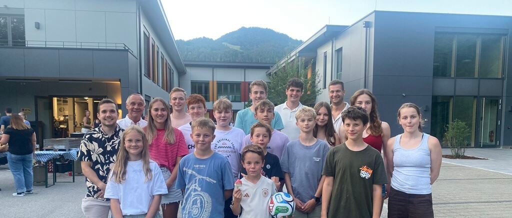 A group of people, including adults and children, are posing for a photo in front of a building with a mountain in the background. Some are holding a soccer ball.