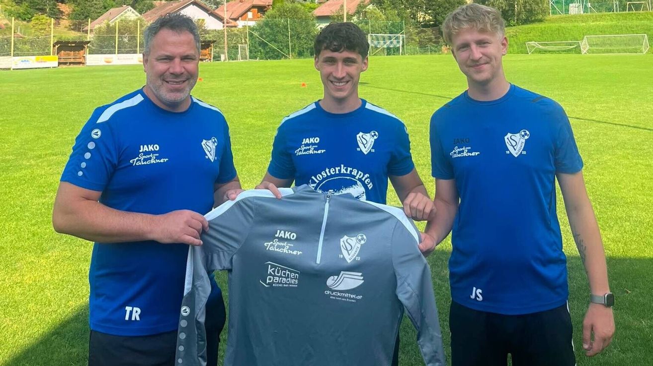 Three men in blue jerseys stand on a soccer field, one holding a gray jersey with white text and logos. They are smiling and posing for a photo.