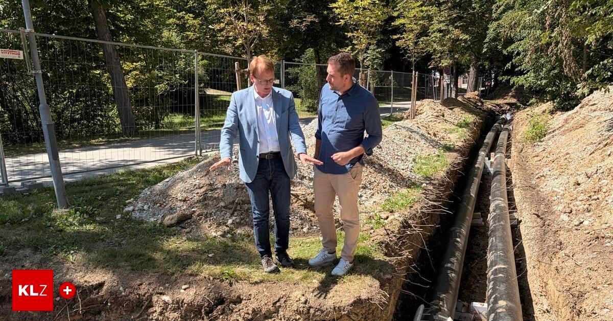 Two men are standing in a park, one wearing a blue blazer and the other a blue shirt, discussing something near a construction site with a fence and trees in the background.
