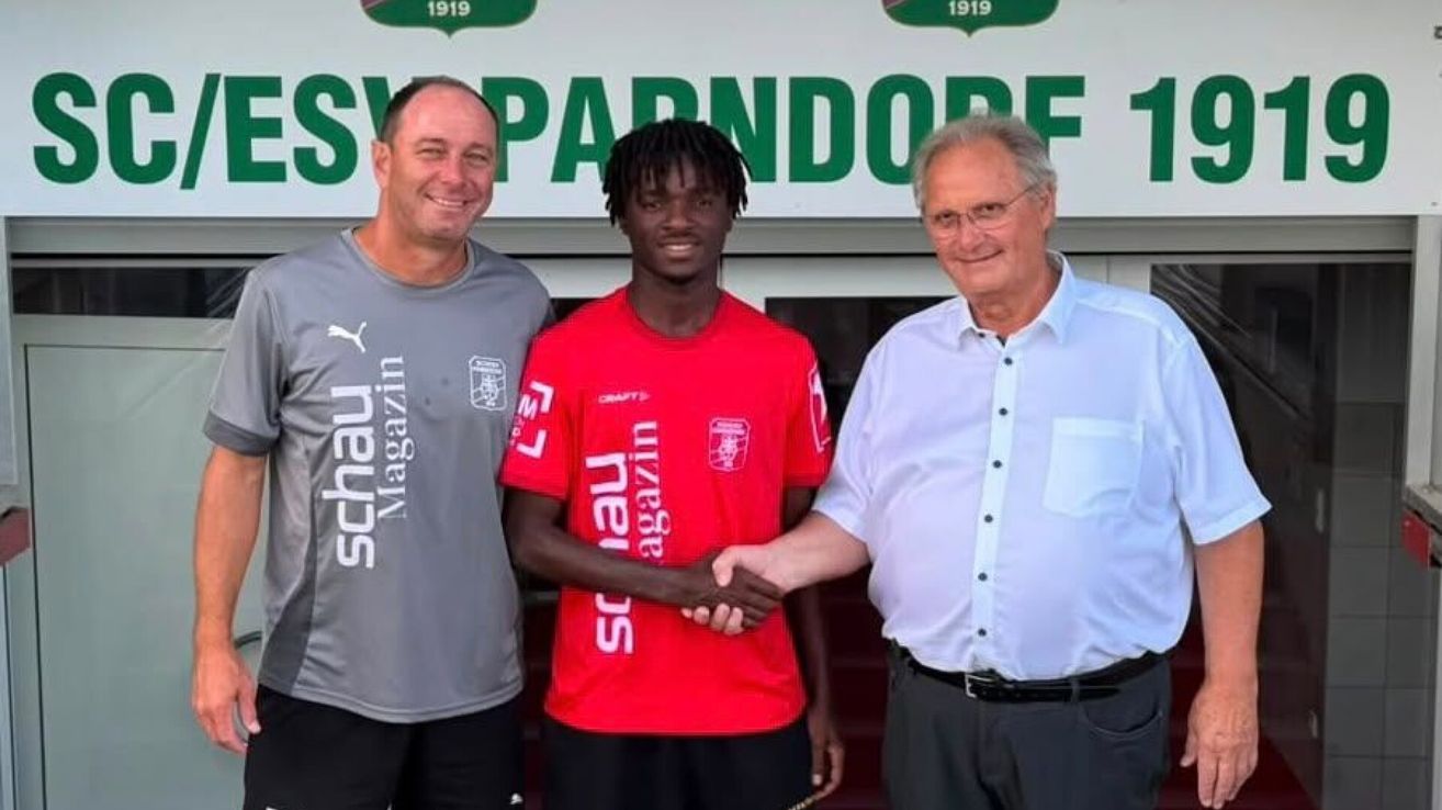 Drei Männer stehen vor einem Schild, das 'SPARENDORF 1919' besagt. Der Mann in der Mitte trägt ein rotes Fußballtrikot mit dem Wort 'Schalke' darauf. Der Mann auf der linken Seite trägt ein graues Shirt mit dem Wort 'Puma' darauf. Der Mann auf der rechten Seite trägt ein weißes Hemd und eine Brille.
