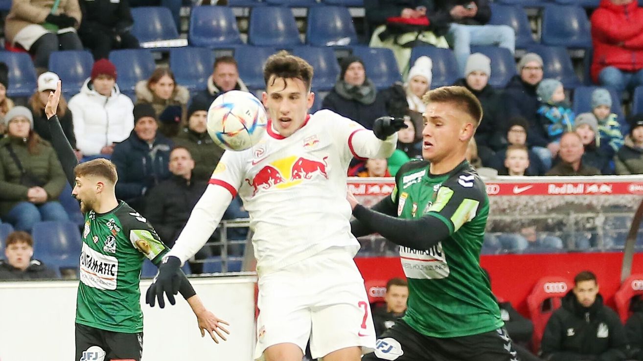 A soccer player in a white uniform with a red bull logo attempts to head the ball while another player in green tries to block him. Spectators in winter clothing watch from the stands.