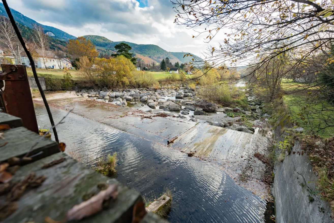 A serene view of a river with rocks and greenery, mountains in the background under a cloudy sky.