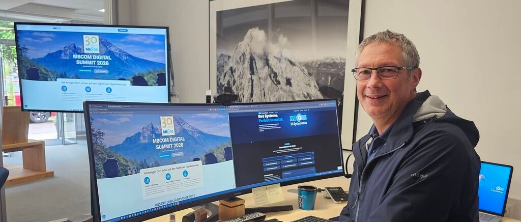 A man sits at a desk with three computer monitors displaying the MBCOM Digital Summit 2026 website. He wears glasses and a blue jacket. Behind him is a large photo of a mountain.