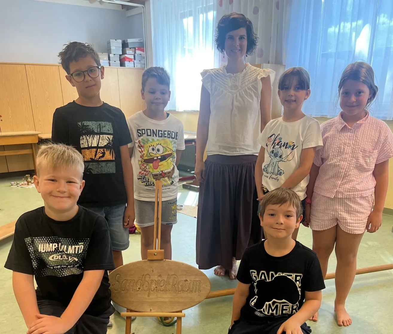 A group of children and a woman stand in a room, smiling for a photo. The children wear casual clothes, and the woman wears a white blouse. A wooden easel with a sign reading 'Sandspielraum' is in the foreground.