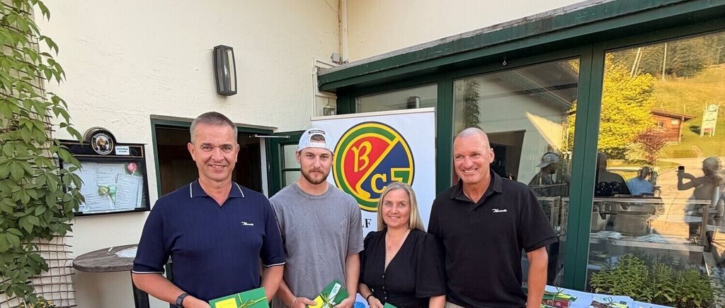 Four individuals stand outside a building with a logo, smiling and holding green gift boxes. The man on the left wears a navy polo, the middle man a gray t-shirt, and the two on the right wear black polos.