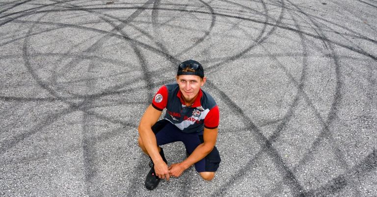 A man in a racing uniform kneels on a gravel surface with tire marks, smiling for the camera.
