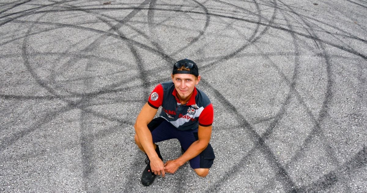 A man in a racing uniform kneels on a gravel surface with tire marks, smiling for the camera.