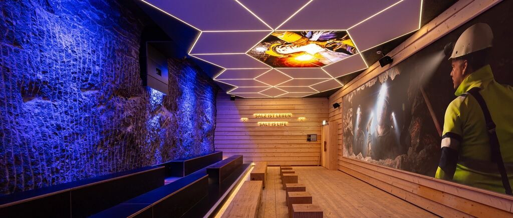 An indoor cinema with wooden benches and a large screen showing a person in a cave with a hammer. The ceiling has hexagonal lights and a large sign that says 'Salt is Life'.