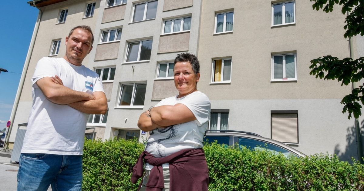 A man and a woman stand in front of a multi-story apartment building with their arms crossed, smiling for the camera.