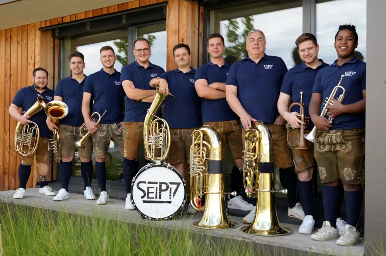 A group of men dressed in traditional Bavarian attire, including blue shirts and brown shorts, are standing together holding brass instruments. They are positioned in front of a building with glass windows.