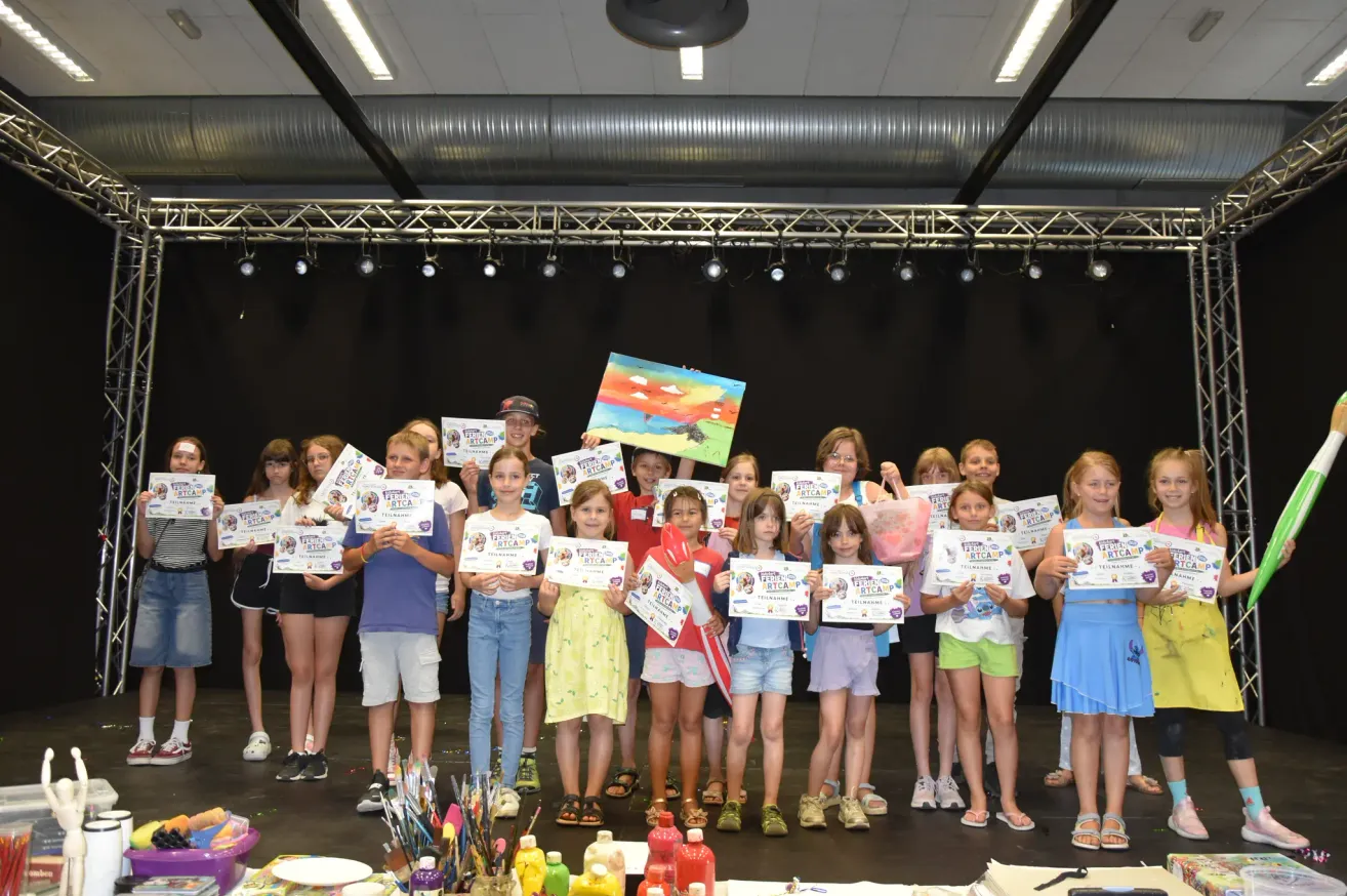 Eine Gruppe von Kindern, die Urkunden halten und auf einer Bühne stehen, einige mit Farbe an den Händen. Die Bühne ist mit einem schwarzen Hintergrund und Scheinwerfern dekoriert.