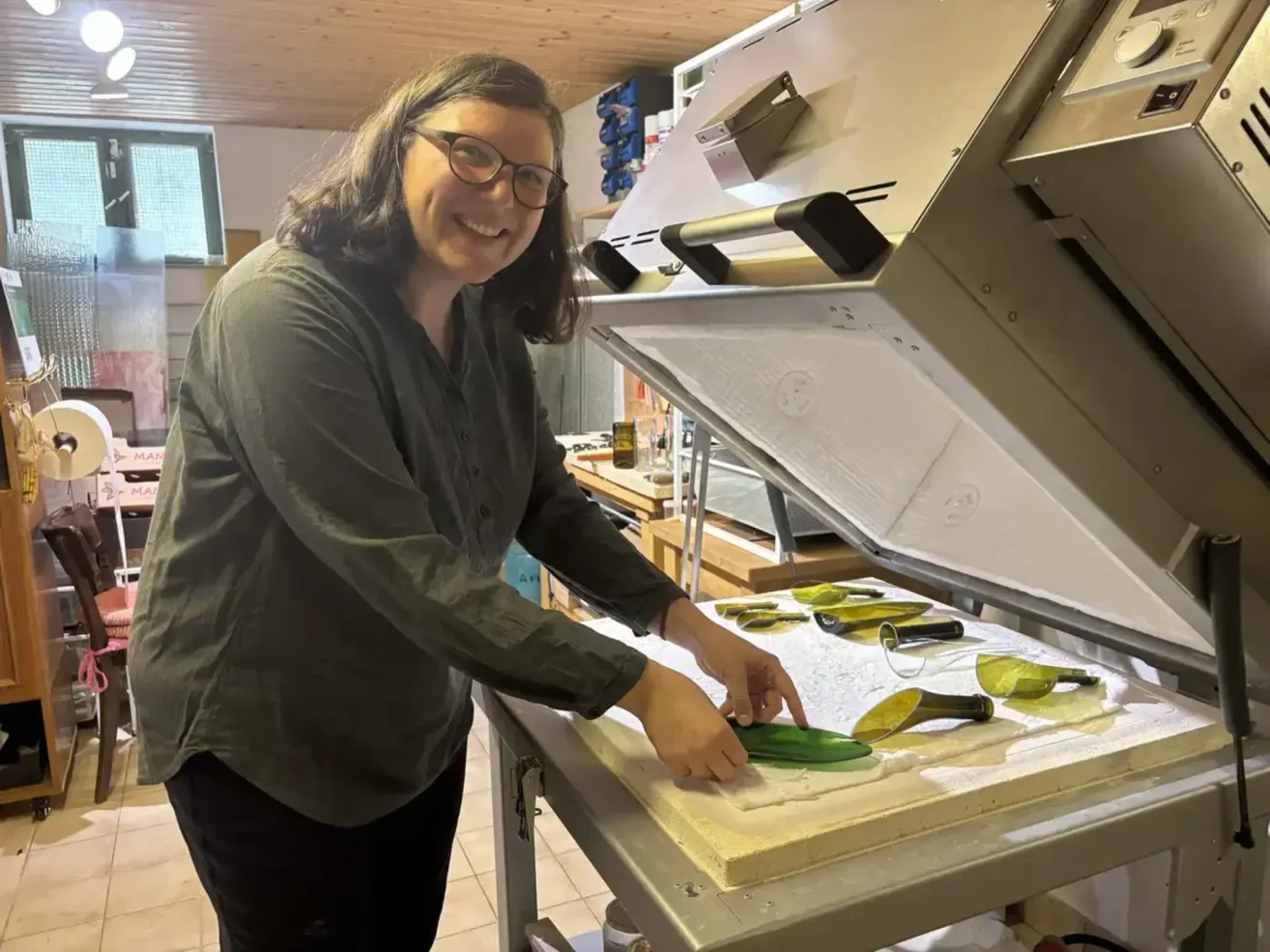 A woman wearing glasses and a green shirt is working on a glass craft in a workshop. She is smiling and holding a green glass piece.