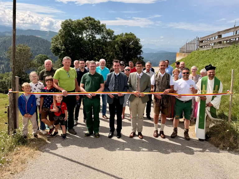 Eine Gruppe von Menschen in traditioneller Kleidung steht in einer Reihe, hält ein Band und posiert für ein Foto auf einer Straße mit einer malerischen Kulisse.