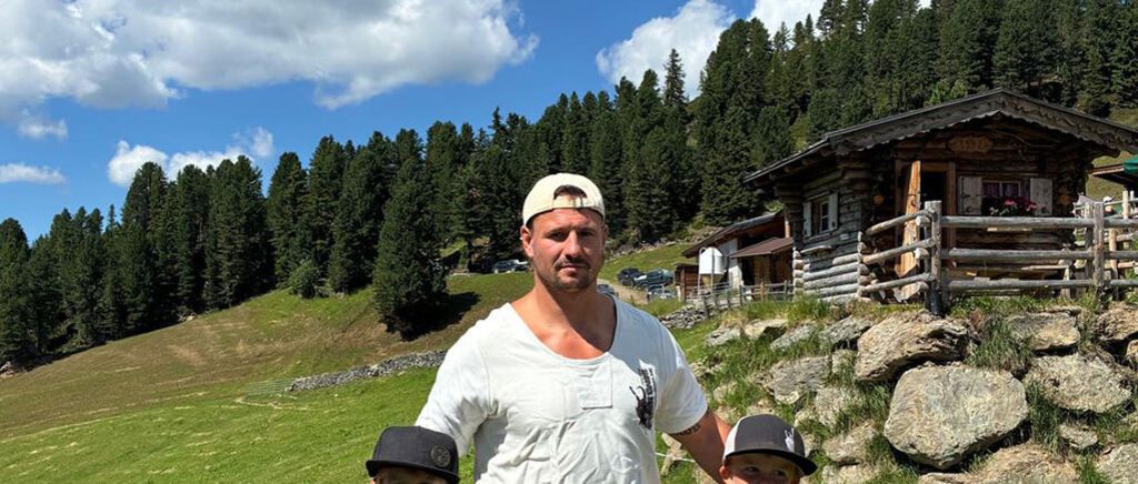 A man in a white t-shirt and a white cap stands with two children in front of a wooden house in a mountainous area.