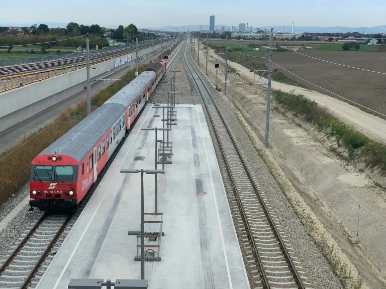 A red train is on the tracks, moving towards a platform. The platform is empty, and the train is at the station. The background shows a city skyline.
