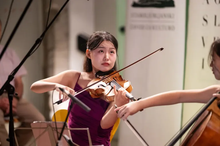 A young woman plays the violin in a concert, wearing a purple dress and holding the violin with both hands. Another person is playing a violin behind her.