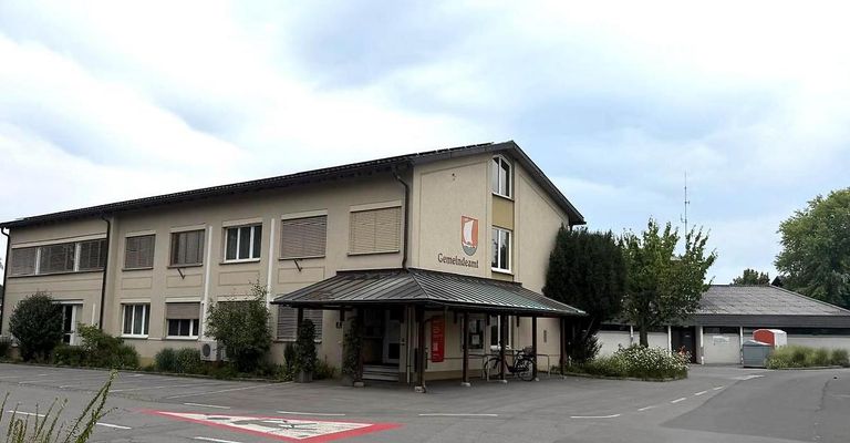 A building with a gray exterior and a black roof, featuring a sign reading 'Gemeindeamt'. A bicycle is parked under the canopy in front of the entrance. The building is surrounded by greenery and a parking lot.