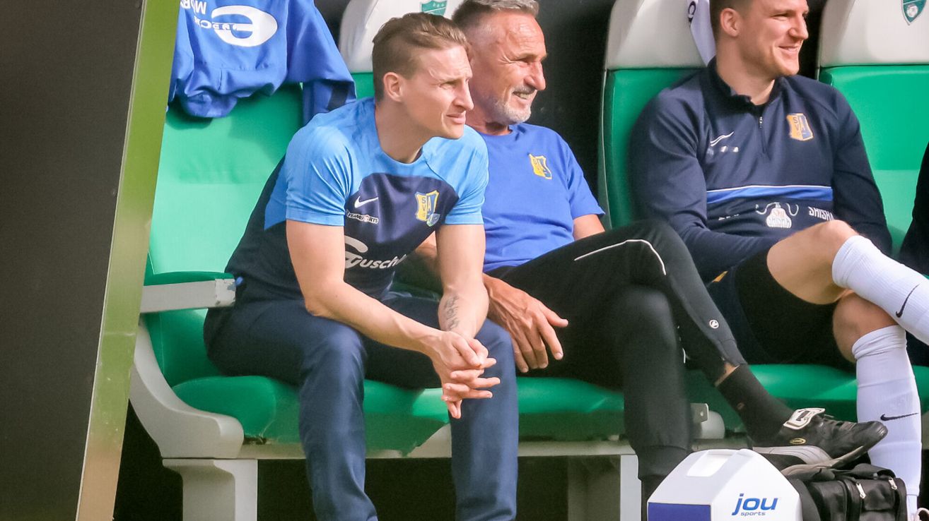 Two men in blue jerseys sit on a bench, one with a tattoo on his arm, the other with a beard and a yellow logo on his shirt.