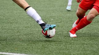 A soccer player in white is about to kick a soccer ball, while another player in red is defending.