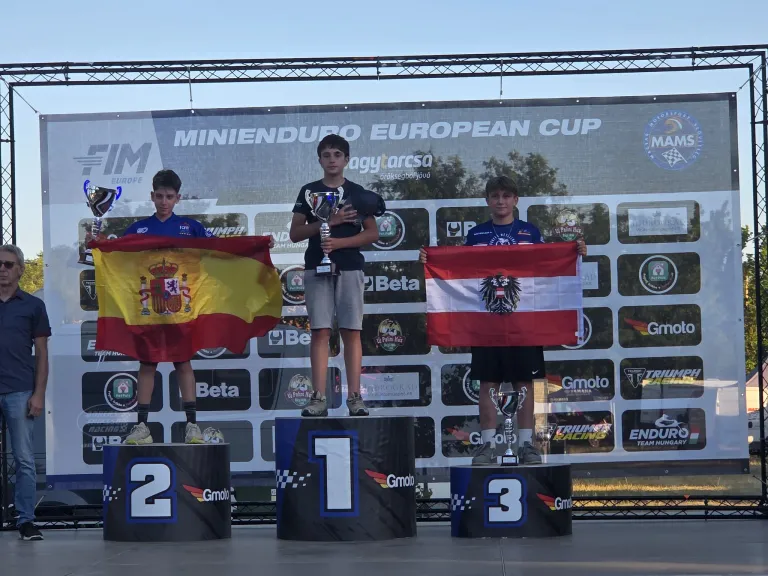 Three young boys stand on a podium holding trophies and flags, with the first-place boy holding an Austrian flag, the second-place boy holding a Spanish flag, and the third-place boy holding a German flag. Behind them is a large banner with the words 'MINIENDURO EUROPEAN CUP'.