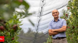A man in a light blue shirt and brown pants stands with his arms crossed in an orchard. He is smiling and appears to be posing for a photo. Behind him, there is a net and apple trees. The background is blurry.