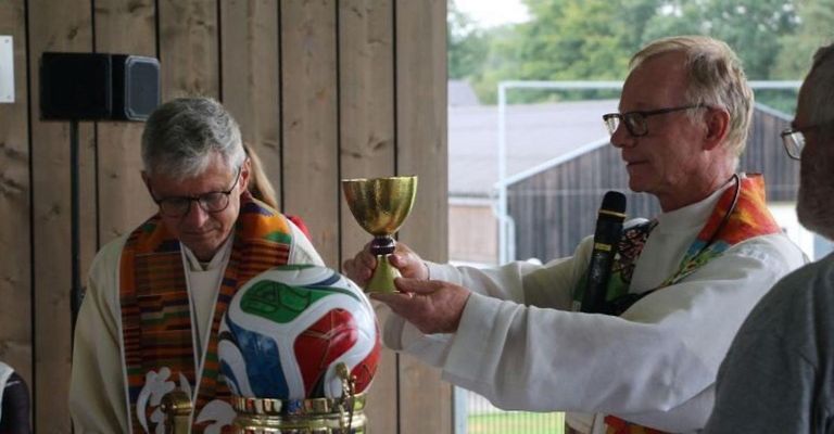 Two men in clerical attire hold a golden chalice and a colorful soccer ball, likely during a religious ceremony.