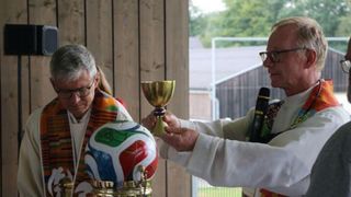 Two men in clerical attire hold a golden chalice and a colorful soccer ball, likely during a religious ceremony.